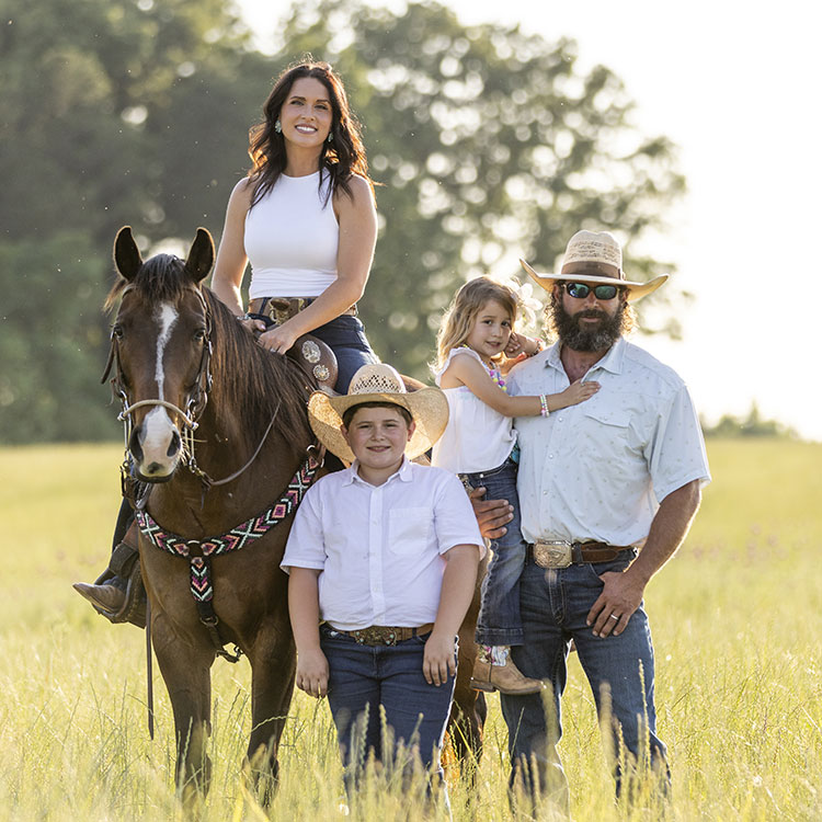 The Smith Family, Kip, Kalie, Tarver, and Cecillia, dressed in Cowboy attire, in a field, while Kalie with black hair is riding a brown horse. 